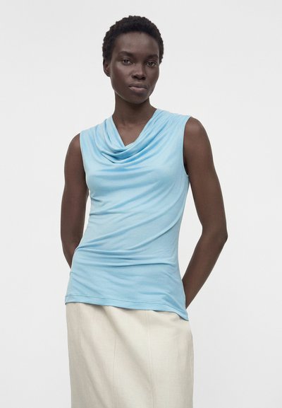 Γυναίκα με κοντά μαλλιά που φοράει ένα αμάνικο γαλάζιο τοπ με πτυχωτό ύφασμα και μια μπεζ φούστα, στέκεται μπροστά σε ένα απλό φωτεινό φόντο.