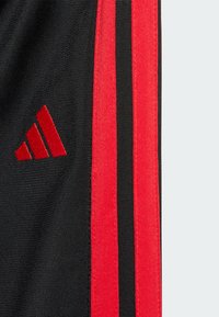 Pantalones cortos deportivos negros con rayas verticales rojas y un logo bordado en rojo. La tela es texturizada y duradera para un uso activo.