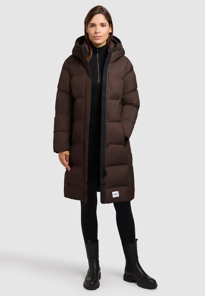 Manteau matelassé marron avec capuche, doté d'une fermeture éclair et de boutons, texture moelleuse et un patch logo près de l'ourlet, associé à un pantalon noir et des bottes.