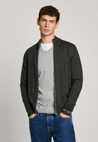 Grå zip-up kofta med ribbade manschetter, lagd över en ljusgrå v-ringad tröja, ihop med blå jeans. Slät textur, modern design.