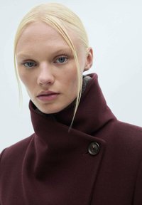 Burgunderfarvet uldfrakke med høj krave, udstyret med en enkelt mørk knap og blød tekstur, båret over en grå indvendig foring.