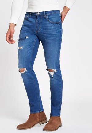 Man in blauwe slim-fit gescheurde spijkerbroek en bruine suède enkellaarsjes, met een gedeeltelijk zichtbare witte trui.