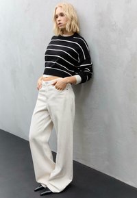 Maglione corto a righe bianche e nere abbinato a jeans bianchi a vita alta e gamba larga, indossato con tacchi neri a punta, davanti a una parete grigia testurizzata.
