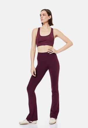 Vrouw staand met hand op heup, draagt bijpassende bordeauxrode sportbeha en uitlopende leggings, witte sneakers tegen een effen achtergrond.