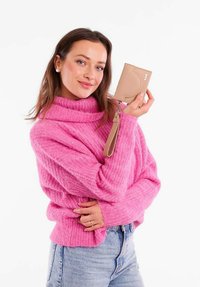 Beige peněženka s zápěstním popruhem, s texturovaným povrchem a třemi dekorativními cvoky, držena osobou v růžovém svetru.