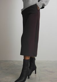 Hoge taille, donkerbordeaux culottes met zijzakken, gecombineerd met zwarte puntige enkellaarsjes met ritssluiting. Gladde stoftextuur.