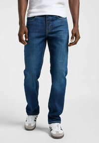 Vaqueros de denim azul con corte recto, que presentan costuras amarillas y un diseño clásico de cinco bolsillos, combinados con zapatillas blancas y grises.