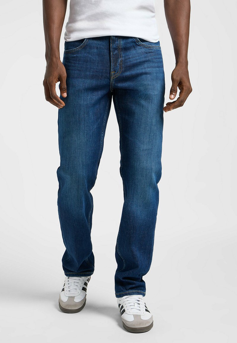 Vaqueros de denim azul con corte recto, que presentan costuras amarillas y un diseño clásico de cinco bolsillos, combinados con zapatillas blancas y grises.