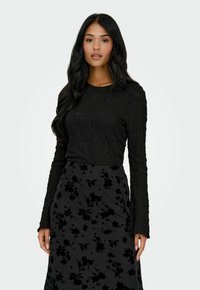 Donna con lunghi capelli neri che indossa un top a maniche lunghe nero dalla texture particolare e una gonna scura con motivi floreali neri, in piedi su uno sfondo semplice.