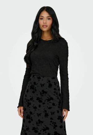 Vrouw met lang zwart haar, gekleed in een gestructureerd zwart shirt met lange mouwen en een donkere rok met zwarte bloemenpatronen, staand tegen een effen achtergrond.