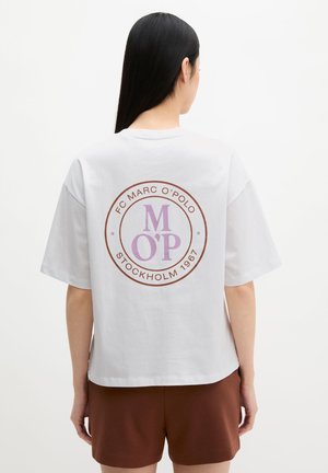 Femme aux longs cheveux noirs porte un t-shirt blanc oversize avec un logo circulaire violet et marron « FC Marc O'Polo Stockholm 1967 », associé à un short marron.