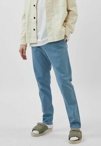 Pantalons en coton bleu clair à coupe droite, associés à une chemise crème et des sandales à glissière vert olive, soulignant une coupe décontractée et un style casual.