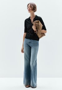 Zwarte gebreide top met korte mouwen en een kraag, gecombineerd met lichtblauwe flared jeans en een beige suède jas in de hand.