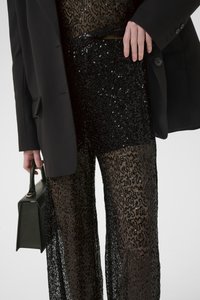 Pantaloni neri a palazzo in paillettes con vita alta e sovrapposizione trasparente testurizzata, abbinati a un blazer nero over-size e a una borsa rettangolare.
