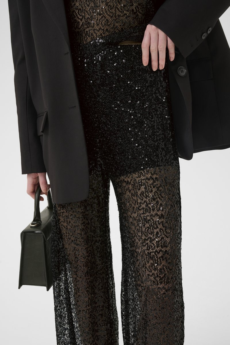 Sorte palietterede højtaljede brede bukser med et gennemsigtigt struktureret overlay, parret med en oversized sort blazer og en rektangulær håndtaske.
