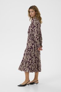 Langes, fließendes Kleid mit burgunderrotem Grundton und beigen Paisleymustern, elastischer Taille und langen Ärmeln. Kombiniert mit schwarzen Kitten-Heels.