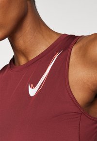 Débardeur bordeaux Nike avec un col rond, arborant un logo blanc et orange clair en haut à gauche. Texture de tissu lisse.