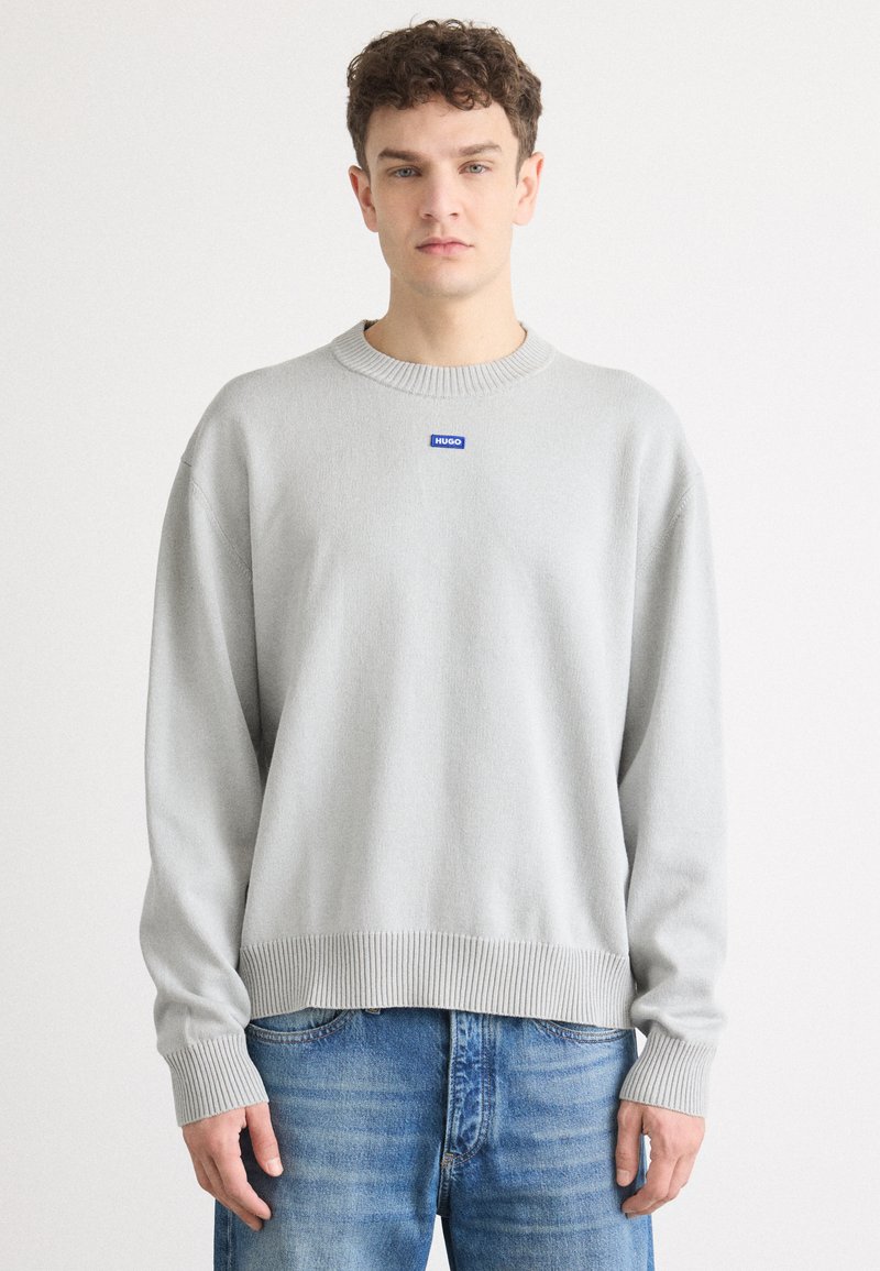 Maglione grigio chiaro a girocollo con orlo e polsini a coste, caratterizzato da un'etichetta con logo blu sul petto, indossato con jeans blu.