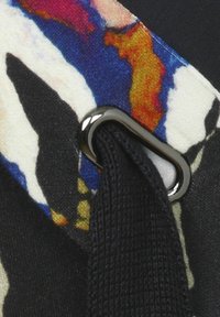 Textilband mit einem farbenfrohen Druck in Orange-, Blau- und Schwarztönen, gehalten von einer metallischen Schlaufe und strukturiertem schwarzem Stoff.