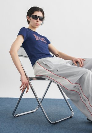 Młoda osoba w ciemnych okularach przeciwsłonecznych, granatowej koszulce Nike oraz jasnoszarych spodniach Nike Shox, siedzi swobodnie na metalowym składanym krześle.