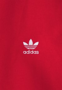Červená bavlněná látka s ribbenou texturou, na které je bílé vyšívané logo Adidas a tři pruhy nad ním.
