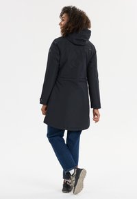 Giacca nera impermeabile con cappuccio, dotata di una cintura in vita e polsini con bottoni. Indossata con jeans blu e calzature nere.