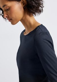 Frau mit kurzen lockigen Haaren, die ein schwarzes Langarmshirt trägt, den Kopf nach unten geneigt vor einem schlichten hellen Hintergrund.