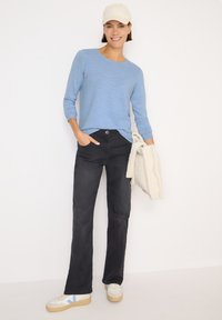 Himmelblaues, strukturiertes Langarmshirt, schwarze gerade Jeans und weiße Sneakers mit blauen Akzenten. Hält eine cremefarbene, flauschige Jacke.