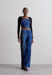 Gombos farmer crop top magas derekú, bő szárú farmernadrággal párosítva; sötét és világos kék részekkel, kontrasztos varrással és fekete hosszú ujjakkal.