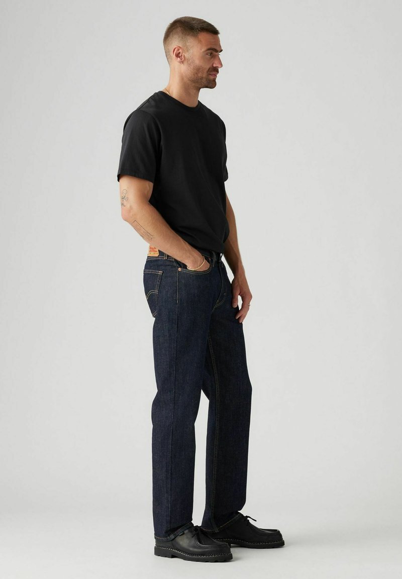 Maglietta nera in cotone abbinata a jeans denim blu scuro. I jeans presentano cuciture gialle e un classico design con 5 tasche. Scarpe nere completano il look.