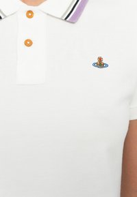 Fehér pólóing, textúrált anyagból készült. Kontrasztos, csíkozott gallérral és hímzett logóval rendelkezik a mellkasán. A gombok kerekek és narancssárgák.