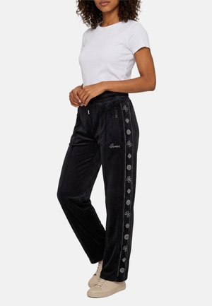 Vrouw met zwarte veloursbroek met patroon zijstrepen en een wit shirt met korte mouwen, staand met handen nabij de taille en beige schoenen.