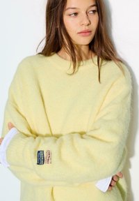 Żółty, puszysty sweter z długimi rękawami i okrągłym dekoltem. Na lewym rękawie znajduje się naszywka, a pod spodem warstwowo założona jest biała koszula.