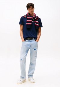 Tamsiai mėlynas marškinėliai, šviesiai mėlyni dilgėliniai džinsai, balti sportbačiai. Raudonas ir tamsiai mėlynas dryžuotas megztas šalikas per pečius.