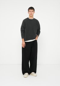 Junger Erwachsener in einem ausgewaschenen schwarzen Langarmshirt, weit geschnittenen schwarzen Hosen und weißen Turnschuhen, der vor einem schlichten weißen Hintergrund steht.