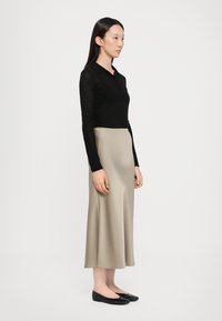 Svart langermet polo skjorte, knappert ved kragen. Beige sateng midi skjørt, glatt tekstur. Svart leiligheter. Modell poserte sidelengs mot en ensfarget bakgrunn.