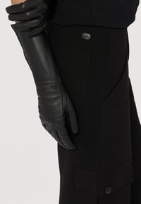 Svarta läderhandskar med en slät yta och skräddarsydd design, visade tillsammans med svarta, vinklade byxor med fickor som har knappdetalj.