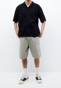 Camicia nera a maniche corte con bottoni e motivo testurizzato, abbinata a pantaloni corti grigi chiari fino al ginocchio e sneakers bianche e nere.