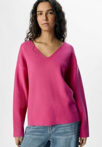 Suéter rosa brillante con escote en V y mangas largas, confeccionado en una tela suave. Combinado con jeans de mezclilla azul claro. Diseño simple y relajado.