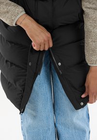 Svart vadderad väst med dragkedja och knappstängning, bärs över ljusblå jeans av denim. Texturerade ärmar syns i bilden.