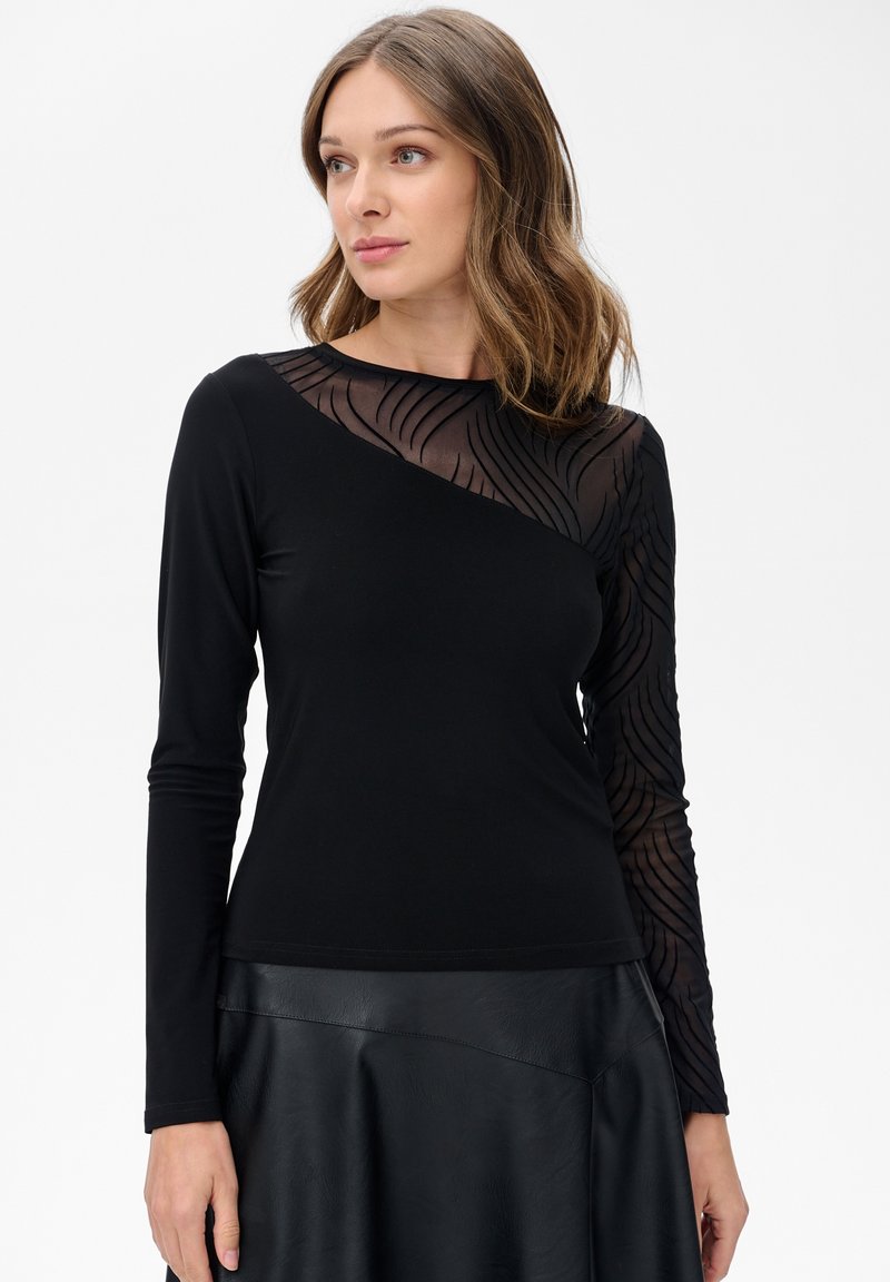 Schwarzes Langarmoberteil mit einem transparenten Mesh-Einsatz mit Wellenmuster auf einer Schulter, kombiniert mit einem satinähnlichen schwarzen Rock.