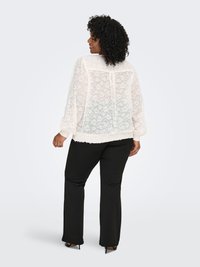 Genomskinlig vit blus med blommigt spetsmönster, långa ärmar och elastiska ärmslut; kombinerad med svarta byxor och skor med klack.