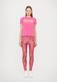T-shirt in cotone rosa con logo "VERSACE JEANS COUTURE", abbinato a leggings rosa con un colorato motivo floreale e sneakers bianche.
