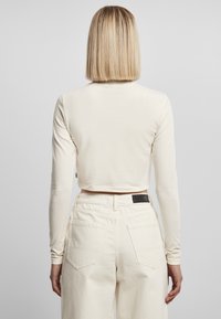 Cremiges Langarm-Crop-Top mit Rundhalsausschnitt, kombiniert mit hoch taillierten hellbeigen Hosen mit Gesäßtaschen und einem schwarzen Lederpatch.