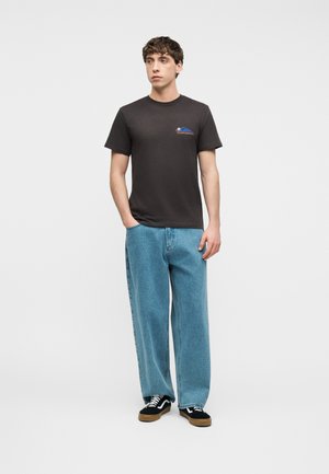 Νέος άνδρας που φοράει μαύρο τυπωμένο t-shirt, φαρδιά ανοιχτό μπλε τζιν και μαύρα αθλητικά παπούτσια με λευκά κορδόνια, στέκεται μπροστά σε απλό φόντο.