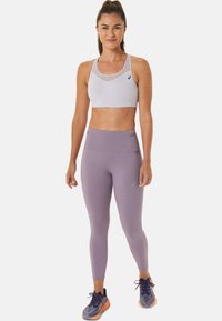 Hellgrauer Sport-BH mit einem Mesh-Design, kombiniert mit high-waist lavendelfarbenen Leggings, die über ein dezentes Logo verfügen. Modell trägt Sport Schuhe.