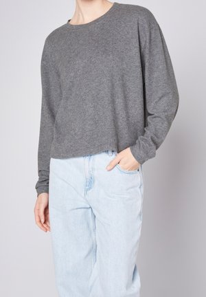 Henkilö, jolla on yllään väljälahkeinen harmaa pitkähiha neulepaita ja vaaleansiniset korkeavyötäröiset farkut, toinen käsi taskussa.