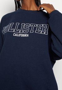 Granatowa bluza z bawełnianej mieszanki, z wyraźnym białym napisem "HOLLISTER CALIFORNIA" na przodzie, z raglanowymi rękawami.