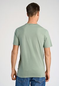 Mężczyzna z krótkimi brązowymi włosami, ubrany w jasnozieloną koszulkę z krótkim rękawem i niebieskie dżinsy, stoi tyłem na jednolitym tle.