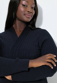 Selvsikker kvinde med langt mørkt hår iført en marineblå ribstrikket sweater står med armene krydsede foran en hvid baggrund.